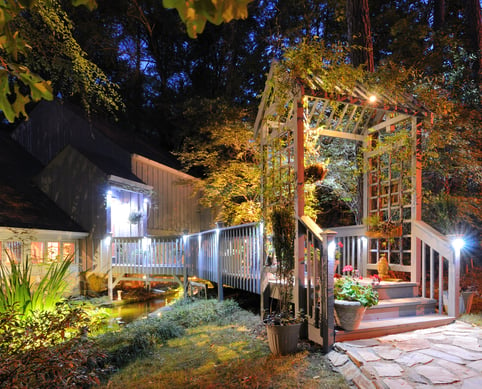 House exterior with porch walkway over a pond and lighting in the woods